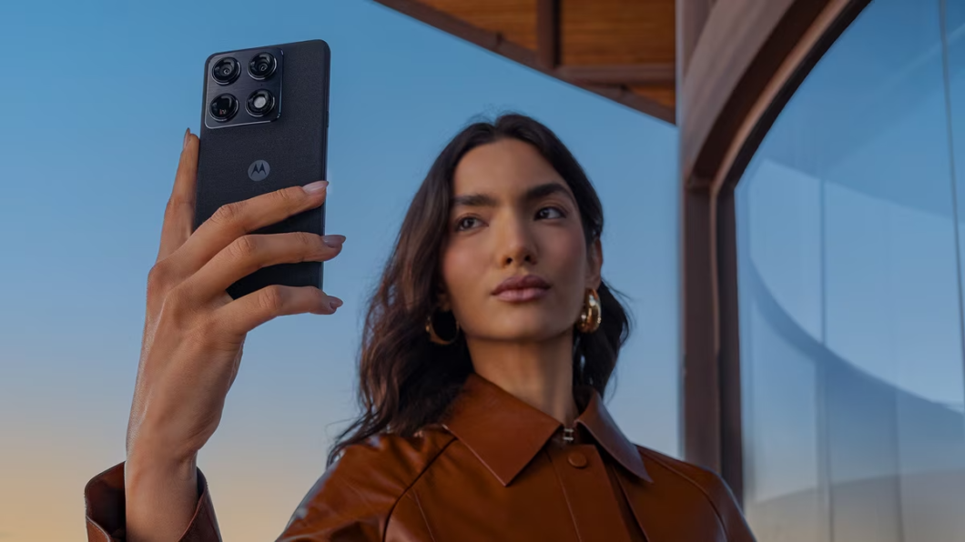 Mulher segura um smartphone Motorola Signature com a câmera traseira voltada para frente, como se estivesse tirando uma selfie. O aparelho tem acabamento escuro, textura que simula tecido e módulo quadrado com quatro câmeras. Ao fundo, há uma estrutura de madeira e vidro, com céu azul ao entardecer.