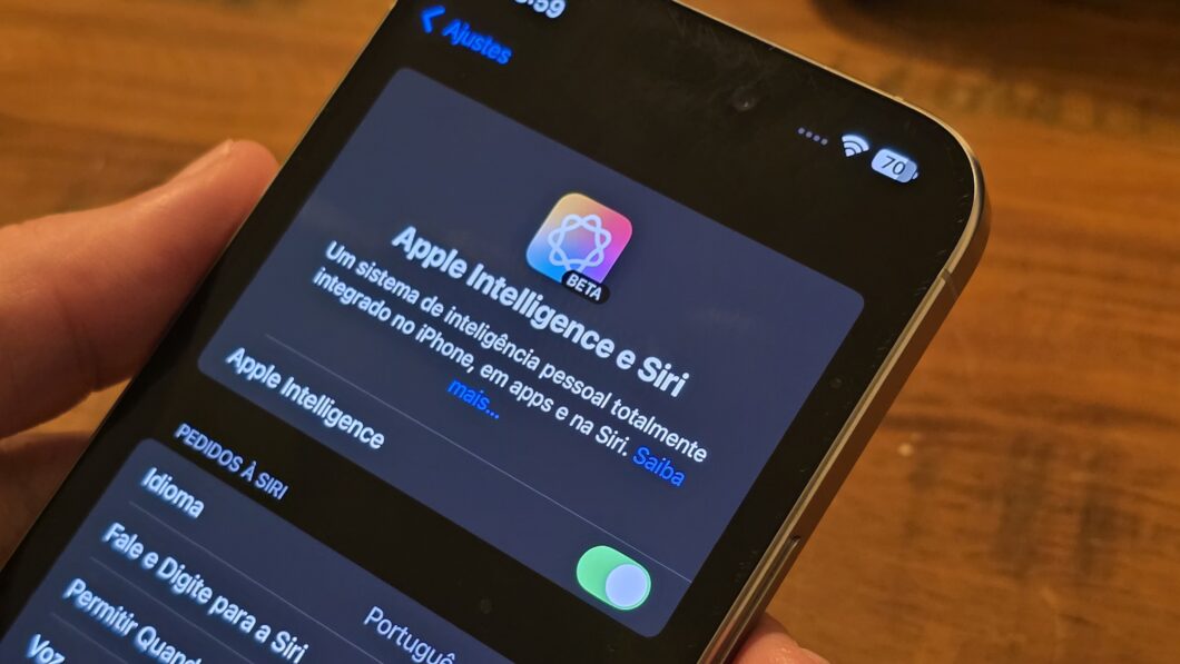 Um iPhone sendo segurado por uma mão exibe a tela de configurações da Apple Intelligence e Siri, com um design escuro. O texto informa que se trata de um sistema de inteligência pessoal integrado ao iPhone e à Siri, ainda em fase beta. O botão de ativação está ligado. A interface está em português, e no canto superior direito, há ícones de Wi-Fi e bateria indicando 70% de carga. O fundo da imagem tem uma superfície de madeira desfocada.