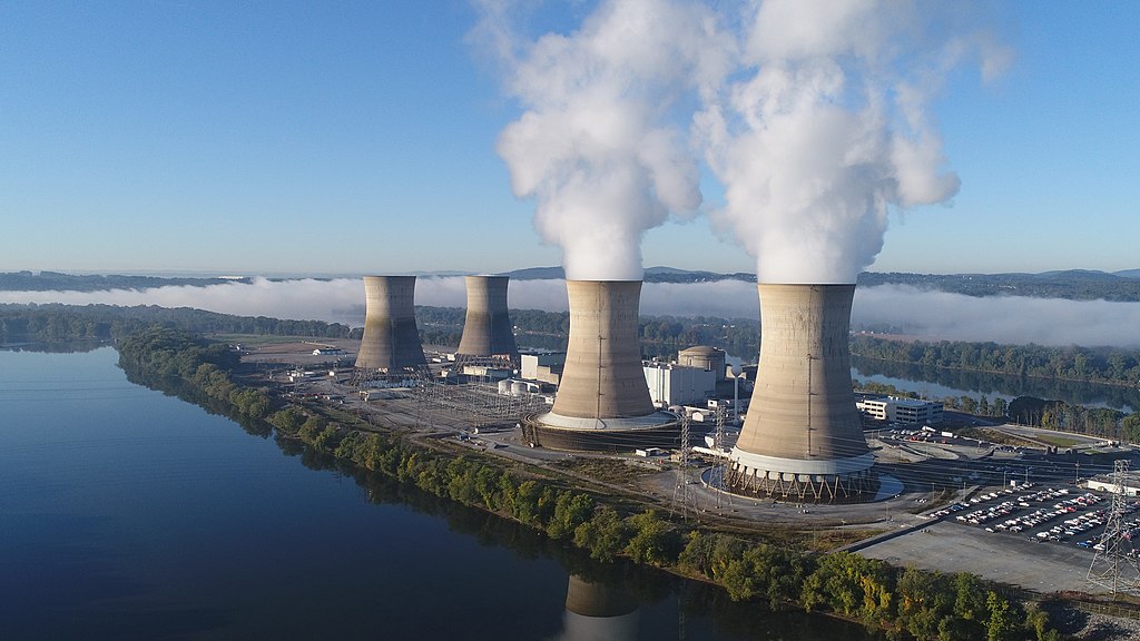 Fotografia aérea da Usina Nuclear de Three Mile Island, na Pensilvânia. A usina está situada em uma ilha cercada por um rio calmo, sob um céu azul claro. No centro, destacam-se quatro grandes torres de resfriamento em formato de hiperboloide; as duas à direita emitem densas nuvens de vapor branco, enquanto as duas à esquerda estão inativas. Ao fundo, uma névoa baixa cobre a vegetação das margens. Em primeiro plano, vê-se um estacionamento e instalações elétricas.