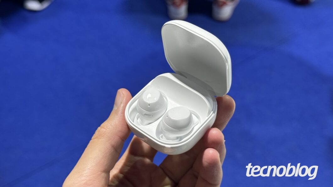 Uma mão segura um estojo de fones de ouvido sem fio de cor branca, que está aberto e revela dois fones também brancos. O logotipo 