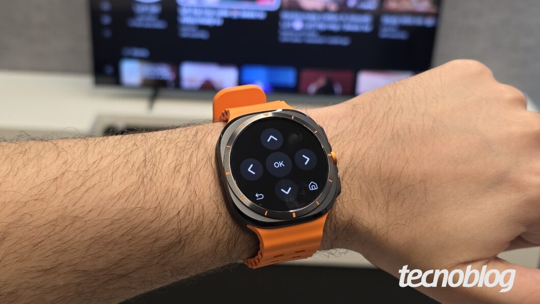 Braço com relógio smartwatch de pulseira laranja, tela exibe controles direcionais com botões 