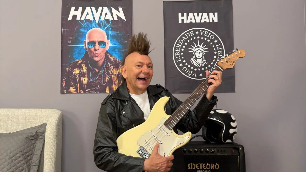 empresário dono da Havan, Luciano Hang, vestido com uma jaqueta de couro preta, segurando uma guitarra bege nas mãos com um cabelo moicano